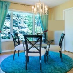A bright dining room featuring a round blue rug, a black table with white chairs, and a chandelier, with a view of green trees through large windows.