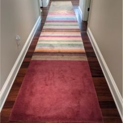 A long, multicolored striped rug laid on a dark wooden hallway floor, bordered by white walls and baseboards.